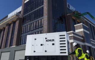 Mobile vs. Stationary Industrial Generators: Which Type Best Fits Your Business Power Requirements | 7 | Long Island Power Systems Two construction workers in safety vests connecting hoses to a large, white, towable Kohler mobile generator outside a modern brick and glass building under construction.